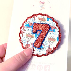 May include: A circular scalloped badge featuring a red glitter border and a central number seven in red glitter with a blue outline. The background pattern displays comic book style illustrations of spider webs and the word POW in yellow.