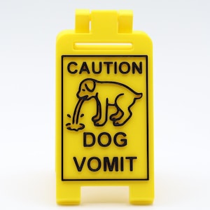 May include: A bright yellow caution sign with black text and a graphic of a dog vomiting. The sign reads "CAUTION DOG VOMIT" in bold, capital letters. The sign is made of plastic and has a rectangular shape.