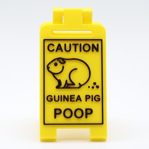 May include: A yellow caution sign with black text and a black silhouette of a guinea pig. The sign reads "CAUTION GUINEA PIG POOP."