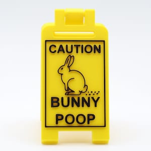 Peut inclure: Un panneau de signalisation jaune avec un lapin noir et le texte "ATTENTION CROTTES DE LAPIN".