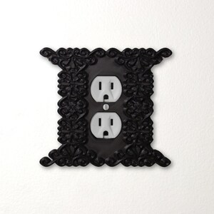 Pu&ograve; includere: Copertura decorativa nera per presa elettrica con design floreale. La copertura ha due prese elettriche standard con prese bianche. La cornice ornata aggiunge un tocco di eleganza a qualsiasi stanza. Montato su una parete bianca.