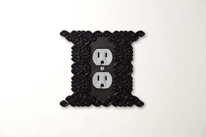 Pu&ograve; includere: Copertura decorativa nera per presa elettrica con design floreale. La copertura ha due prese elettriche standard con prese bianche. La cornice ornata aggiunge un tocco di eleganza a qualsiasi stanza. Montato su una parete bianca.