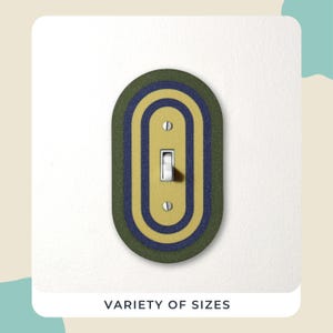 May include: An oval light switch cover with a layered design in olive green, navy blue, and yellow. The switch is white, and the text "VARIETY OF SIZES" is at the bottom. The cover is mounted on a white wall.
