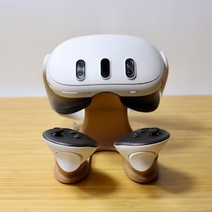 May include: A white virtual reality headset with two black controllers, all resting on a wooden stand.