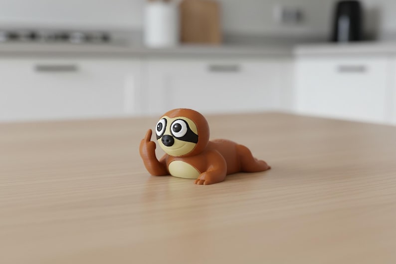 May include: A brown sloth figurine with a black eye mask and large, expressive eyes. The sloth is lying down and giving the middle finger. The figurine is on a light brown wooden surface.