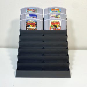May include: A black plastic storage stand for Nintendo 64 game cartridges. The stand has 12 slots and is stacked in a tiered design. The stand is filled with various game cartridges, including "Quest 64", "Diddy Kong Racing", "Pokemon Stadium", and "Super Smash Bros."