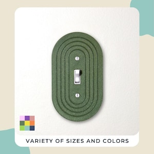 May include: A green, oval-shaped light switch cover with a series of concentric lines. The switch is white, and the cover is mounted on a white wall. The text "VARIETY OF SIZES AND COLORS" is at the bottom.