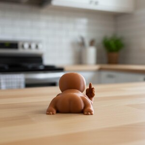 May include: A brown figurine of a baby lying on its stomach, with its rear end facing the viewer and its middle finger raised. The figurine is on a light-colored wooden surface, with a kitchen in the background.