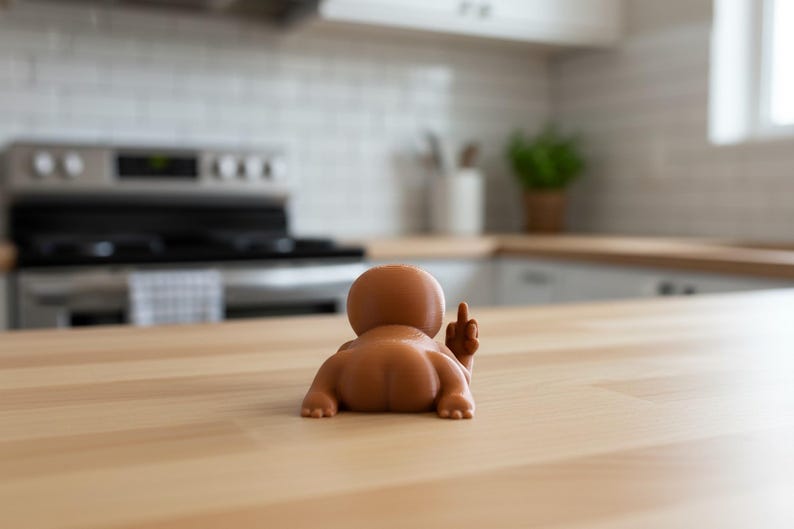 May include: A brown figurine of a baby lying on its stomach, with its rear end facing the viewer and its middle finger raised. The figurine is on a light-colored wooden surface, with a kitchen in the background.
