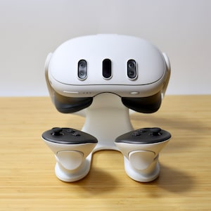 May include: A white virtual reality headset with two black controllers on a white stand. The headset has three black cameras on the front.