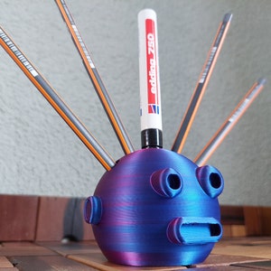 Könnte beinhalten: Ein blauer und lila 3D-gedruckter Stiftehalter in Form eines Gesichts mit Löchern für Stifte und einen Marker. Der Marker ist weiß mit schwarzer Schrift und trägt die Aufschrift "edding 750".