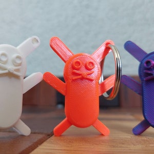 May include: Three 3D printed keychains shaped like cartoon bunnies. The bunnies are white, orange, and purple. The orange bunny has a silver key ring attached.