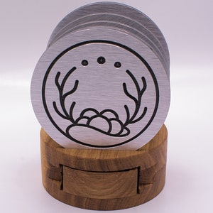 May include: Set of six silver metal coasters with a black outline design of a stylized antler and a cluster of berries. The coasters are stacked and displayed on a wooden stand.