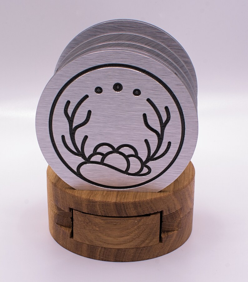 May include: Set of six silver metal coasters with a black outline design of a stylized antler and a cluster of berries. The coasters are stacked and displayed on a wooden stand.