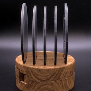 May include: Five round, black metal coasters are stacked on a wooden stand. The stand is made of light brown wood with a natural grain pattern.