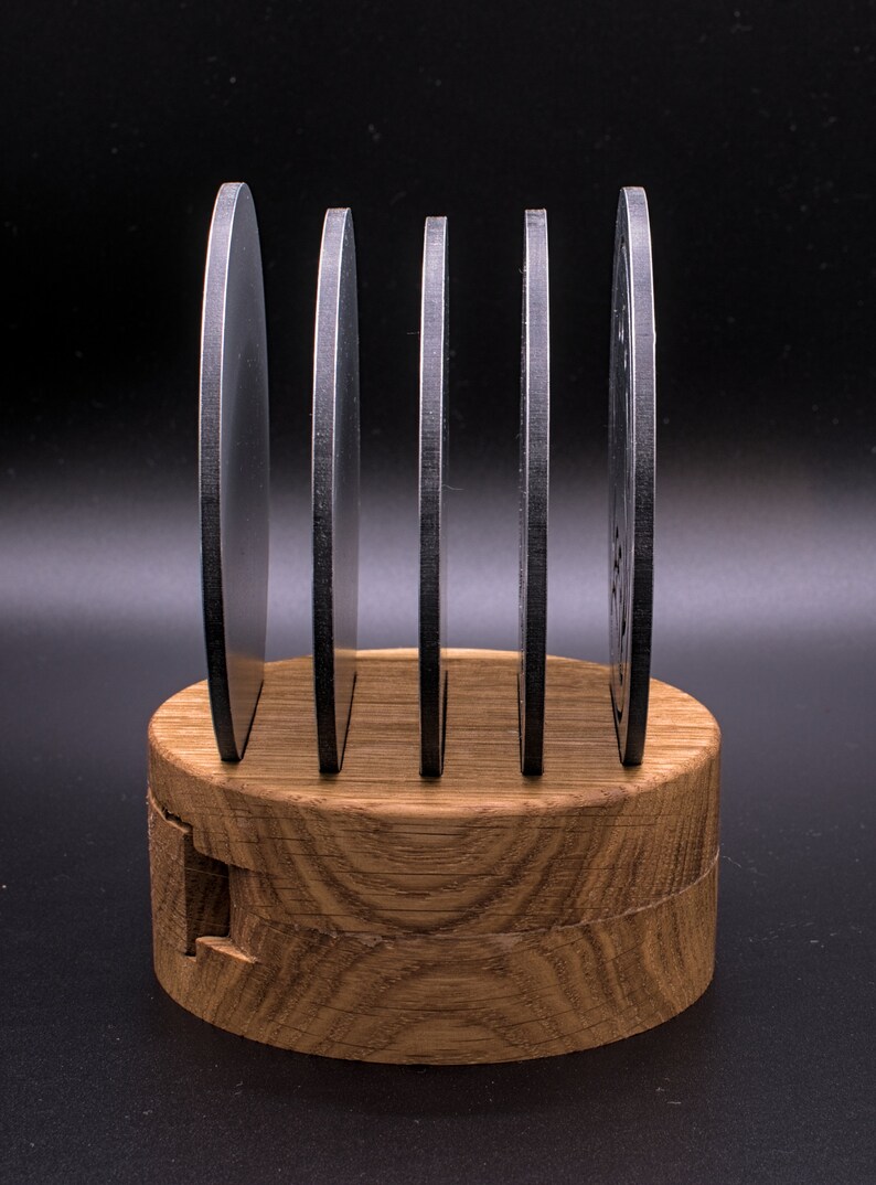 May include: Five round, black metal coasters are stacked on a wooden stand. The stand is made of light brown wood with a natural grain pattern.
