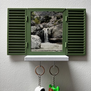 May include: A wall-mounted key holder featuring a rectangular frame with olive green shutters and a central image of a waterfall cascading over rocks. A small white shelf is attached below the frame, with two metal rings hanging from the bottom.