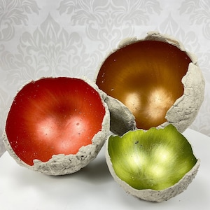 Könnte beinhalten: Drei dekorative Betonschalen mit einer rauen, strukturierten Außenseite. Die Schalen sind in verschiedenen Farben lackiert: orange, grün und gold.