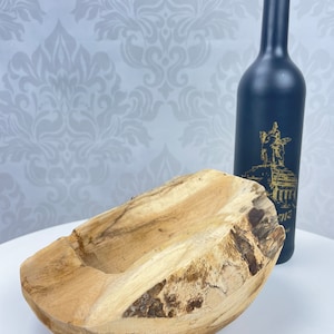 May include: A natural wood bowl with a rough, textured exterior and a smooth interior. A dark blue bottle with a gold design is behind the bowl. The bowl is a light brown color and has a unique, organic shape.