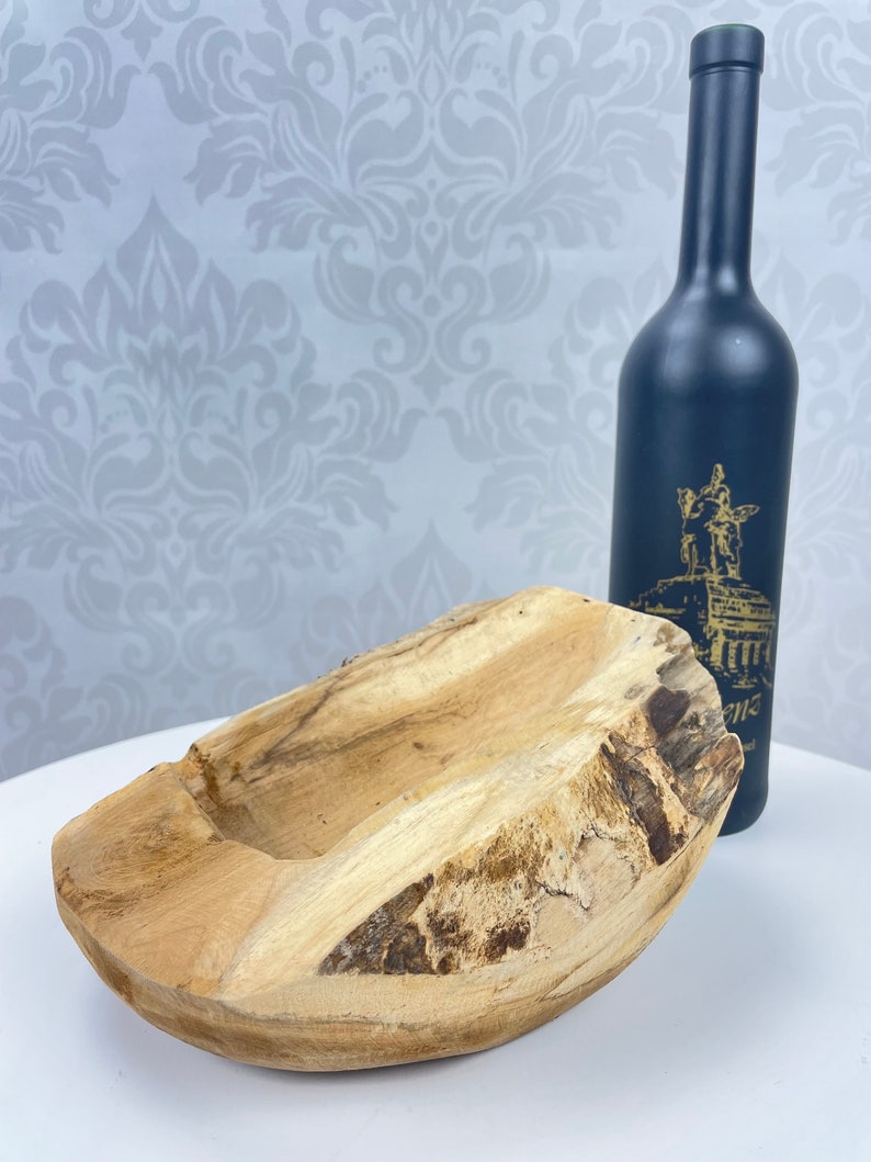 May include: A natural wood bowl with a rough, textured exterior and a smooth interior. A dark blue bottle with a gold design is behind the bowl. The bowl is a light brown color and has a unique, organic shape.
