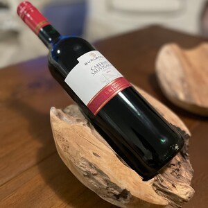 May include: A dark red wine bottle with a red and white label, resting in a natural wood holder. The label reads "BURLWOOD Cabernet Sauvignon". The bottle is angled, showcasing the wine's color and the wood's texture. A decorative piece for wine enthusiasts.