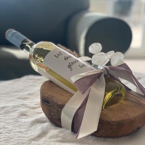 May include: A clear glass wine bottle with a light blue cap, a white label with black text, and a decorative bow. The bottle rests on a wooden base and is tied with a white and lavender ribbon. The text on the label is blurry.