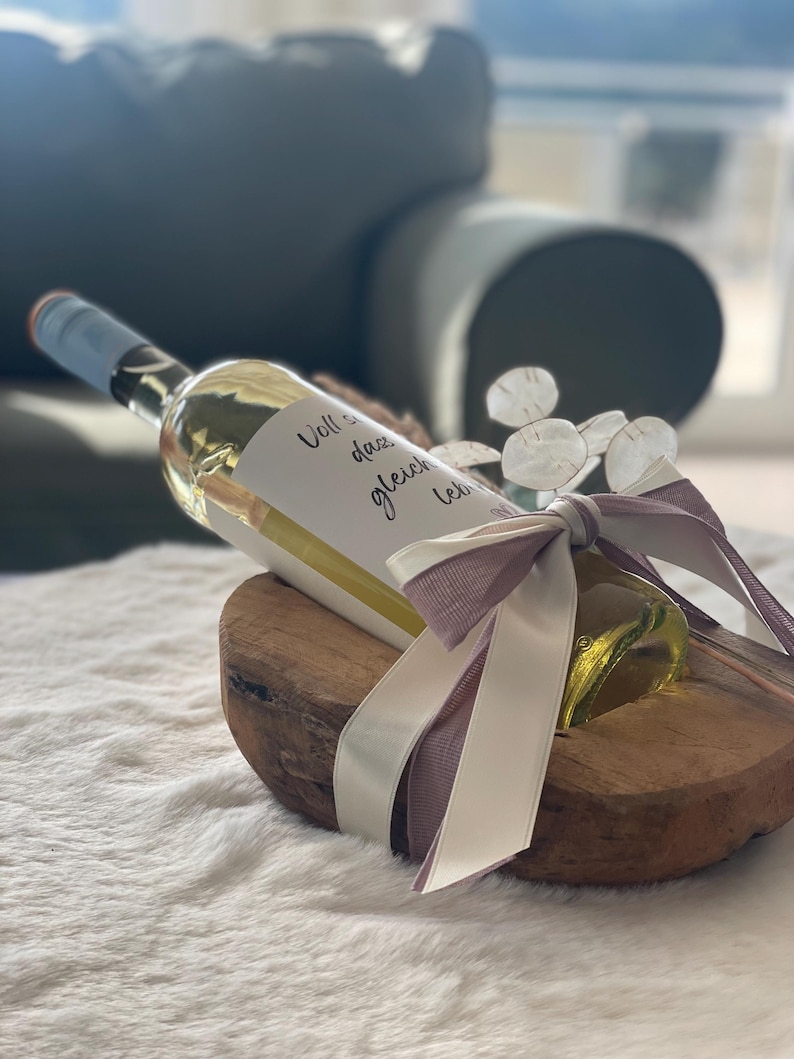 May include: A clear glass wine bottle with a light blue cap, a white label with black text, and a decorative bow. The bottle rests on a wooden base and is tied with a white and lavender ribbon. The text on the label is blurry.
