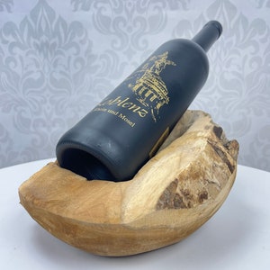 May include: A dark bottle of wine rests in a natural wood wine holder. The bottle has gold lettering and a gold illustration. The wood holder is light brown and has a curved shape.