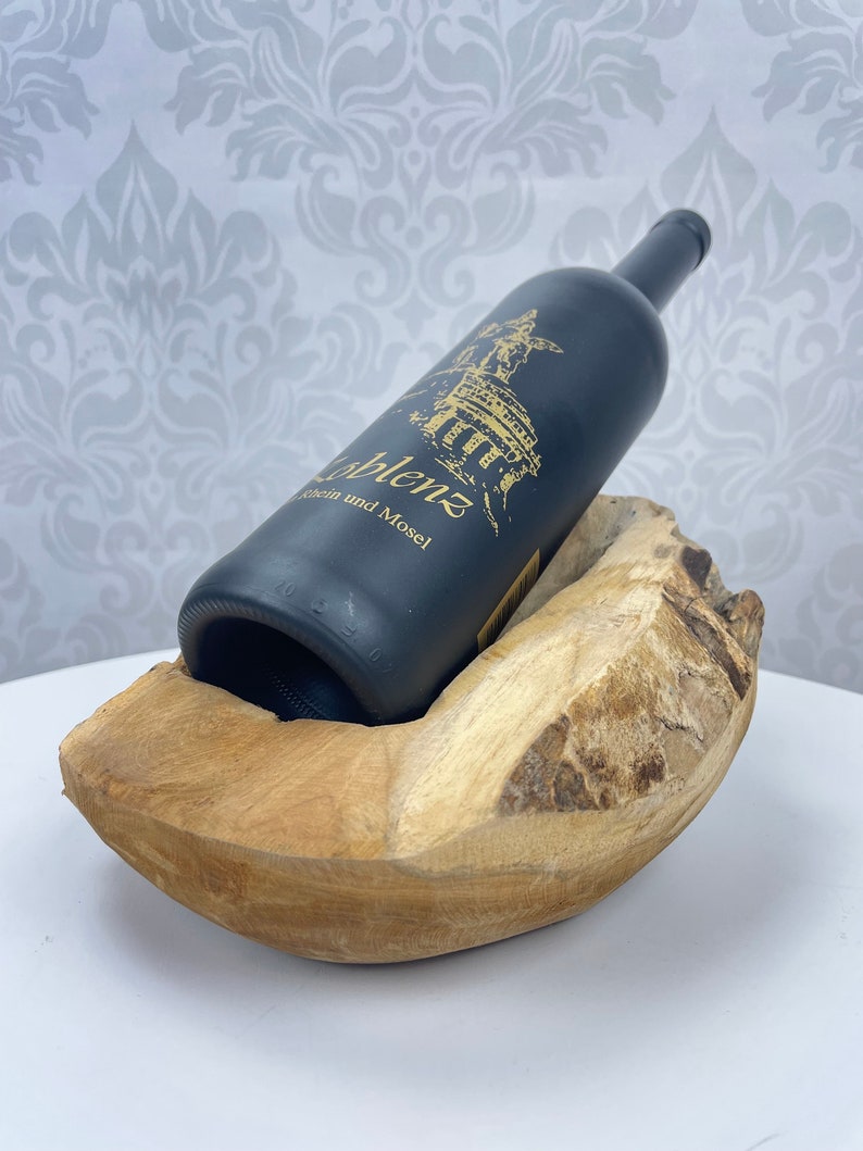 May include: A dark bottle of wine rests in a natural wood wine holder. The bottle has gold lettering and a gold illustration. The wood holder is light brown and has a curved shape.