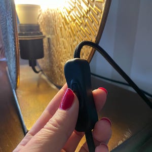 以下が含まれることがあります： 黒い布で覆われた電源コードに取り付けられた黒いインラインロッカースイッチ。スイッチは、金色の質感のある金属製ランプシェードの前に配置されており、背景には点灯した白い電球が見え、照明を提供しています。