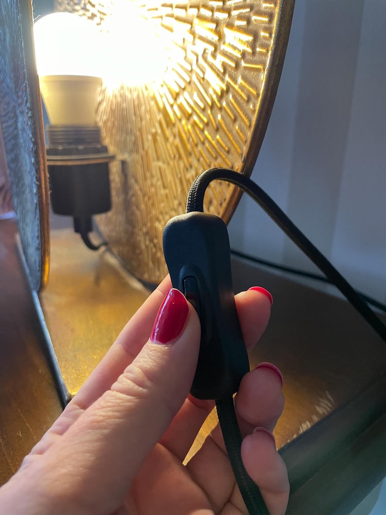 以下が含まれることがあります： 黒い布で覆われた電源コードに取り付けられた黒いインラインロッカースイッチ。スイッチは、金色の質感のある金属製ランプシェードの前に配置されており、背景には点灯した白い電球が見え、照明を提供しています。
