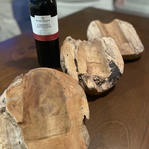 May include: A bottle of Burlwood Cabernet Sauvignon wine next to three rustic wooden bowls. The bowls are irregularly shaped with a natural, light brown color. The wine bottle is dark with a white label.