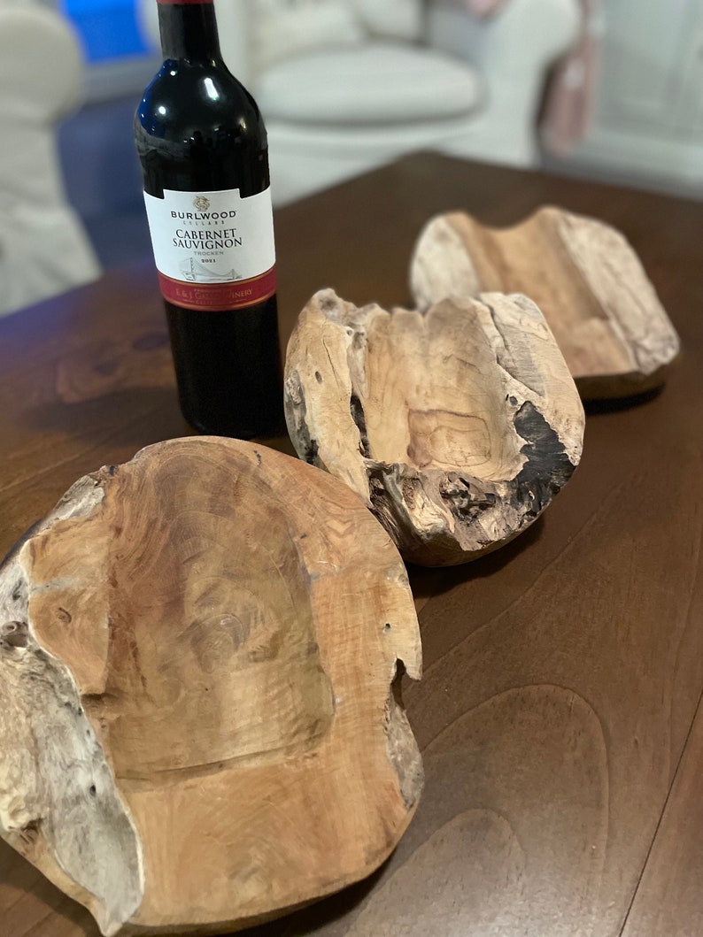 May include: A bottle of Burlwood Cabernet Sauvignon wine next to three rustic wooden bowls. The bowls are irregularly shaped with a natural, light brown color. The wine bottle is dark with a white label.