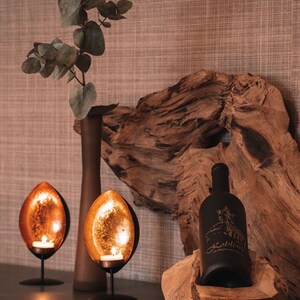 May include: A decorative display featuring two copper-colored, teardrop-shaped candle holders with lit candles. A dark bottle of wine with text is in a wooden bowl. A brown vase and eucalyptus branches complete the arrangement.
