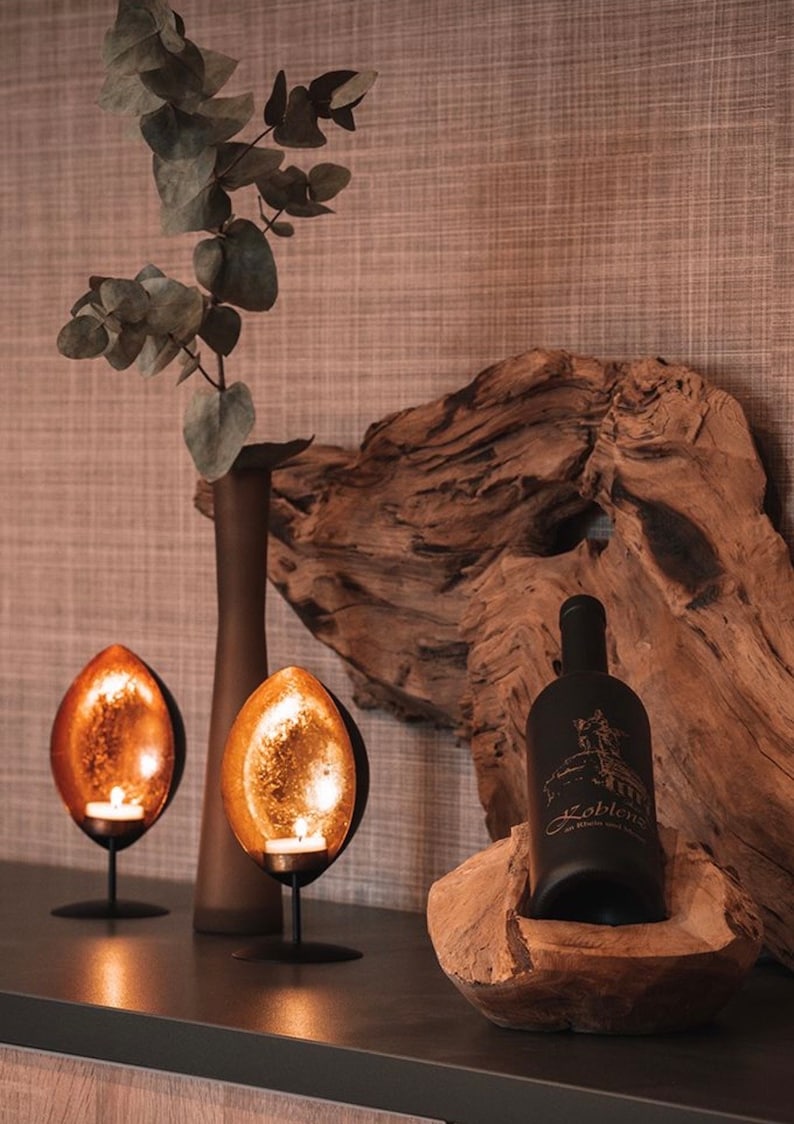 May include: A decorative display featuring two copper-colored, teardrop-shaped candle holders with lit candles. A dark bottle of wine with text is in a wooden bowl. A brown vase and eucalyptus branches complete the arrangement.