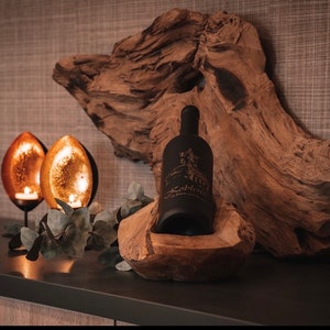 May include: A black wine bottle with a label, nestled in a carved wooden holder, sits on a dark surface. Two candle holders with lit candles and a piece of driftwood complete the rustic decor.