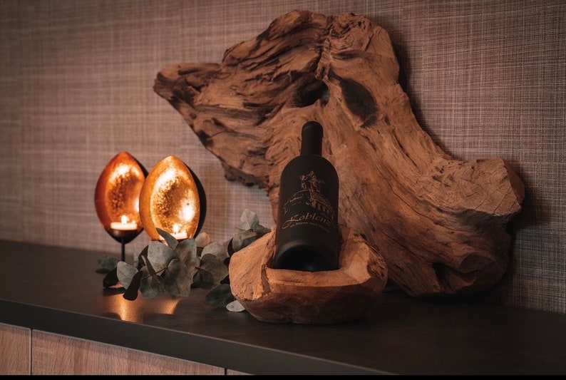 May include: A black wine bottle with a label, nestled in a carved wooden holder, sits on a dark surface. Two candle holders with lit candles and a piece of driftwood complete the rustic decor.