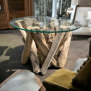 Könnte beinhalten: Ein runder Glastisch mit einer Tischplatte aus Glas steht auf einem Untergestell aus miteinander verwobenen Treibholz-Ästen. Der Tisch befindet sich in einem Wohnzimmer mit einer Couch und anderen Möbeln.