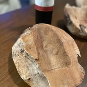 May include: A wooden bowl with a natural, organic shape and a light brown color. The bowl has a smooth, polished interior and a rough, textured exterior. A bottle of red wine is in the background.