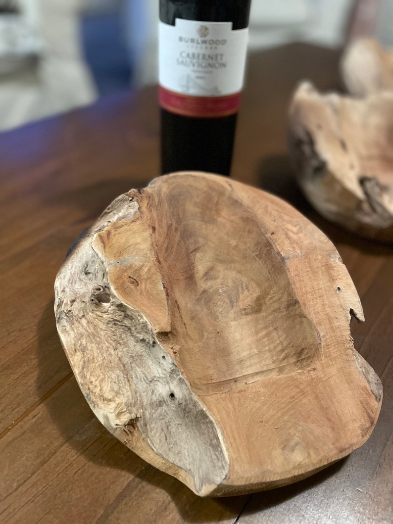 May include: A wooden bowl with a natural, organic shape and a light brown color. The bowl has a smooth, polished interior and a rough, textured exterior. A bottle of red wine is in the background.