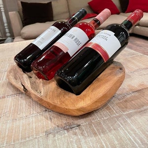 May include: Three wine bottles in a wooden holder. The bottles are filled with red and rose wine. The wooden holder is a natural light brown color and sits on a light-colored wooden table. The background includes a beige couch and red pillows.