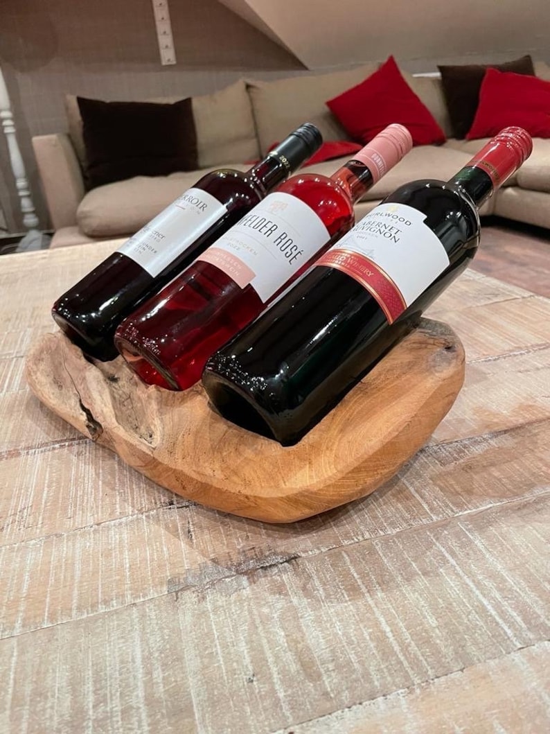 May include: Three wine bottles in a wooden holder. The bottles are filled with red and rose wine. The wooden holder is a natural light brown color and sits on a light-colored wooden table. The background includes a beige couch and red pillows.