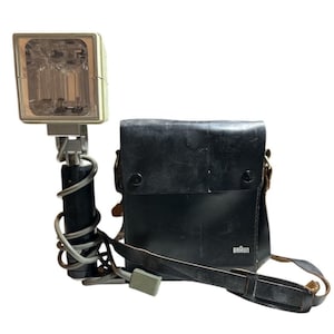 May include: Vintage Braun camera flash with a black handle and a square light head. The flash is connected to a black leather carrying case with a shoulder strap. The case has the Braun logo.
