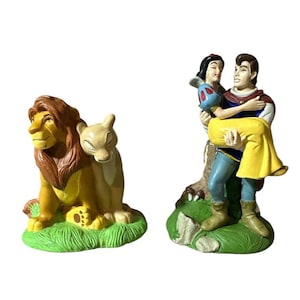 May include: Two ceramic figurines. One depicts a lion and lioness on green grass. The other shows a prince carrying a princess in a yellow dress. The figurines are brightly colored and have a cartoon style.