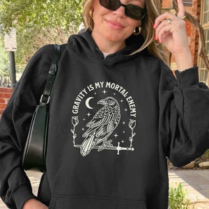 以下が含まれることがあります： 白いカラス、三日月、そして「GRAVITY IS MY MORTAL ENEMY」というテキストがプリントされた黒いフード付きスウェットシャツ。スウェットシャツにはフロントポケットと長袖が付いています。サングラスとライトブルーのジーンズを着用しています。