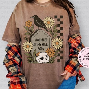 Op de afbeelding: Bruin T-shirt met een Halloween-thema grafisch ontwerp. Het ontwerp toont een grafsteen met de tekst "HAUNTED BY MY OWN BODY", een kraai, bloemen en een schedel. Het shirt wordt gecombineerd met een rood, oranje en zwart geruit flanellen overhemd.