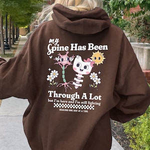 Puede incluir: Sudadera con capucha de color marrón chocolate oscuro con el texto "my Spine Has Been Through A Lot" y un gráfico de un esqueleto de gato, flores y un sol. La sudadera también tiene el texto "but I'm here and I'm still fighting" y "HEALING ONE DAY AT A TIME".