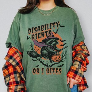 Puede incluir: Camiseta verde salvia con el texto "DISABILITY RIGHTS OR I BITES". El gráfico presenta un mapache montado en una escoba con una calabaza. La camiseta se combina con vaqueros azules y una camisa de franela a cuadros.
