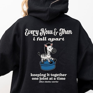 Könnte beinhalten: Schwarzer Kapuzenpullover mit dem Text "Every Now & Then i fall apart" und einem Zeichentrick-Zebra auf einer blauen Plattform. Darunter steht der Text "keeping it together one joint at a time ehlers danlos warrior".