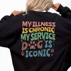 May include: Black sweatshirt featuring the text "MY ILLNESS IS CHRONIC MY SERVICE DOG IS ICONIC" on the back in various colours. The front of the sweatshirt has the text "Chronically Iconic" in colourful letters.